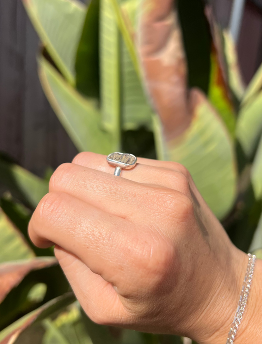 A person's hand wearing a silver ring with a clear rectangular gemstone and a matching silver chain bracelet, shown outdoors in front of green tropical leaves.