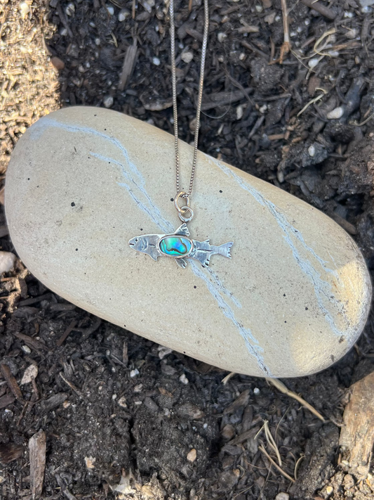 A silver-toned fish pendant necklace with a turquoise-blue oval stone, displayed on a smooth beige stone over dark soil and mulch.