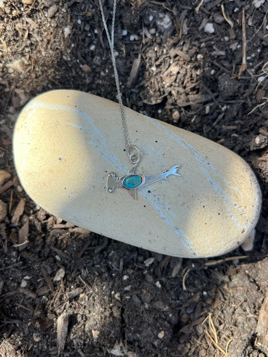 A silver-toned necklace with a turquoise stone pendant is draped over a smooth beige stone placed on dark soil and mulch outdoors.