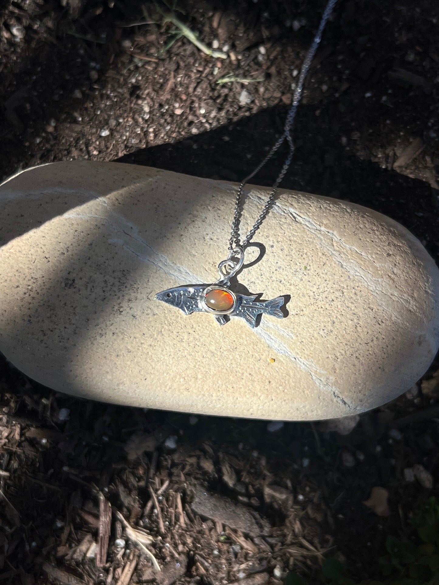 Small fish pendant on a chain lying on a rock with soil and small plants in the background