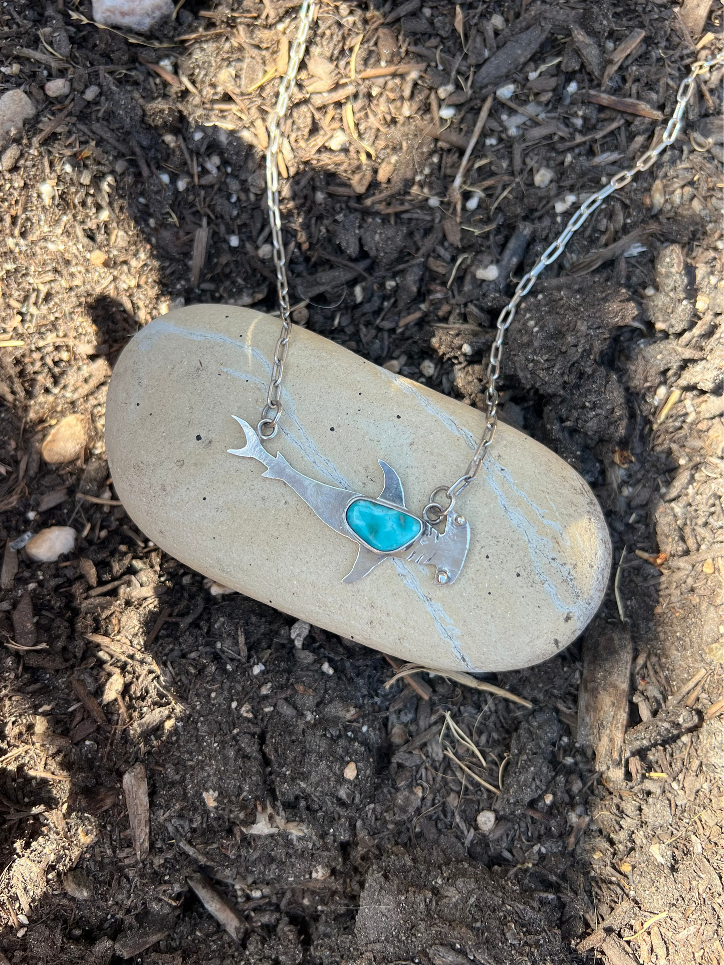 A silver-toned necklace with a shark-shaped pendant and a turquoise heart detail, resting on a smooth beige stone over a bed of dark soil and mulch.