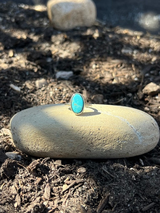 A small oval turquoise ring with a silver band resting on a smooth beige stone outdoors on dark soil.